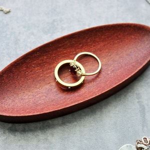 May include: Two gold rings with a red gemstone and clear stones on a dark red wooden ring dish.