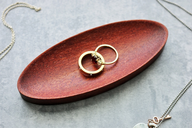 May include: Two gold rings with a red gemstone and clear stones on a dark red wooden ring dish.
