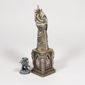 Puede incluir: Una estatua met&aacute;lica detallada de una figura vestida con una t&uacute;nica y una alta tocado, de pie sobre una base decorativa. Una peque&ntilde;a figura gris de un soldado con un arma est&aacute; al lado de la estatua. La estatua mide unos 25 cm de altura.