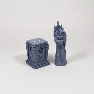 Puede incluir: Dos figuras g&oacute;ticas grises en miniatura. Una es una figura detallada con capucha y t&uacute;nica. La otra es una estructura cuadrada y ornamentada con ventanas arqueadas y detalles esculpidos.