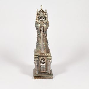 Puede incluir: Una estatua met&aacute;lica detallada de una figura vestida con una t&uacute;nica con las manos juntas, sobre una base decorativa. La estatua tiene un acabado envejecido, similar al bronce, con detalles intrincados en las t&uacute;nicas y la base.