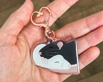 May include: A heart-shaped keyring featuring a black and white rat design. The rats are cuddling and the keyring is made of clear acrylic with a rose gold metal clasp.