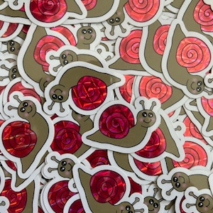May include: A pile of iridescent snail stickers with a red and pink holographic finish. The snails are cartoon-like and have a brown body and a spiral shell.
