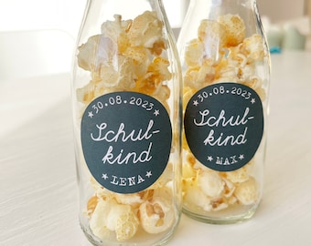 May include: Two glass milk bottles filled with popcorn and decorated with black and white labels. The labels have the text "30.08.2023 Schul-kind" with the names "Lena" and "Max" on each bottle.
