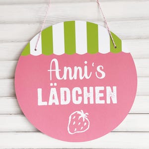 Könnte beinhalten: Rundes, rosa Schild mit dem weißen Text "Anni's Lädchen", gekrönt von einem grün-weiß gestreiften Design. Unten eine weiße Erdbeer-Illustration. Das Schild hängt an einer rosa-weißen Schnur.