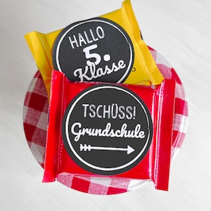 Könnte beinhalten: Draufsicht auf zwei verpackte Süßigkeiten mit runden Etiketten. Die obere Süßigkeit ist gelb mit dem Text "HALLO 5. Klasse" in Weiß. Die untere Süßigkeit ist rot mit dem Text "TSCHÜSS! Grundschule" in Weiß. Die Süßigkeiten liegen auf einem rot-weiß karierten Stoff.