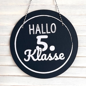 Könnte beinhalten: Rundes, schwarzes Schild mit weißem Text und weißem Rand. Der Text lautet "HALLO 5. Klasse". Das Schild hängt an einer schwarz-weißen Schnur vor einem weißen Holzgrund.