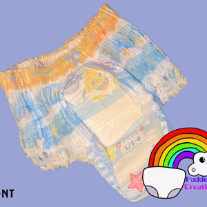 Op de afbeelding: Een baby luier met een kleurrijk ontwerp met stripfiguren. De luier heeft een blauw-witte achtergrond met gele en oranje accenten. De maat is L/5-6. Het woord "FRONT" staat linksonder. Een regenboog- en wolkengrafiek staat rechtsonder.