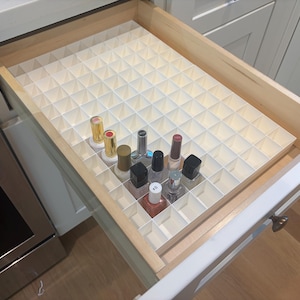 Peut inclure: Un tiroir de cuisine en bois contenant un organisateur à grille en plastique blanc avec de multiples compartiments carrés. Plusieurs petits flacons de vernis à ongles en verre avec des bouchons et contenus variés sont rangés verticalement.