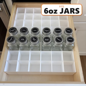 May include: A white plastic drawer organiser with a grid of square compartments. Twelve clear glass spice jars with black circular labels are placed in the front rows. A rectangular sign with an orange border reads FITS 6oz JARS.