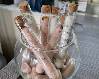 May include: A glass bowl filled with small, smooth, light brown stones. Five glass test tubes with cork tops are placed inside the bowl. The test tubes contain pink and white bath salts and dried herbs.