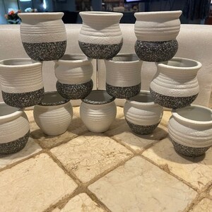 May include: A collection of small, white ceramic pots with a speckled black base. The pots have a textured, ribbed design and are arranged in a group. These decorative pots are suitable for small plants or as home decor.