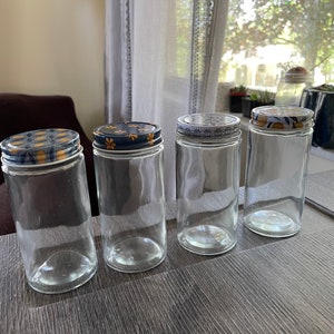 May include: Four clear glass jars with decorative lids. The lids feature floral patterns in blue, yellow, and white. The jars are empty and sit on a wooden surface, suggesting they are for storage or display.