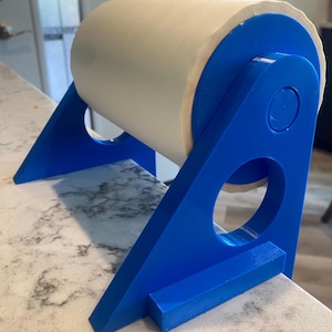 May include: A bright blue plastic paper towel holder featuring a triangular frame with circular cutouts on each side. The holder supports a large roll of white paper towels, which is suspended between the two blue side panels of the stand.