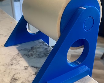 May include: A bright blue plastic paper towel holder featuring a triangular frame with circular cutouts on each side. The holder supports a large roll of white paper towels, which is suspended between the two blue side panels of the stand.