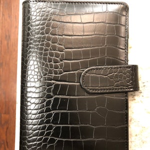May include: Black leather planner with a crocodile embossed pattern and a flap closure.