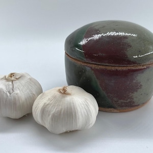 May include: A ceramic garlic keeper with a green, brown, and red glaze. The lid is slightly domed and the base is round. Two whole garlic bulbs are in the foreground.