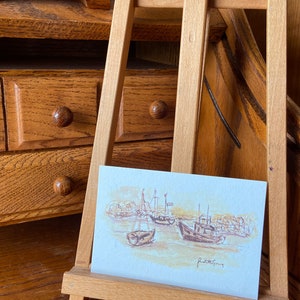 May include: A watercolor painting of boats in a harbor, displayed on a small wooden easel. The artwork features brown and beige tones, depicting several boats and the shoreline. The artist's signature is visible.