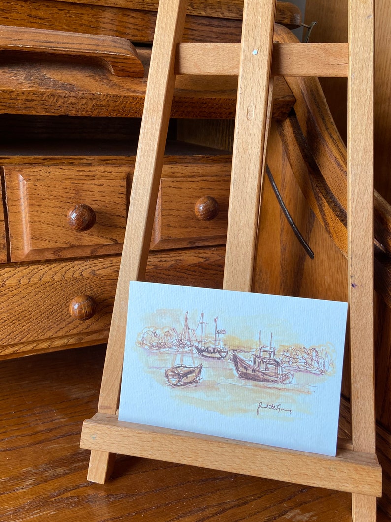 May include: A watercolor painting of boats in a harbor, displayed on a small wooden easel. The artwork features brown and beige tones, depicting several boats and the shoreline. The artist's signature is visible.