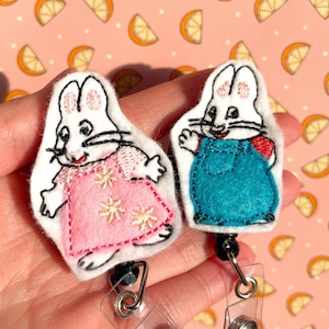 May include: Two embroidered bunny character badge reels with retractable cords. The bunny on the left is wearing a pink dress with white stars and the bunny on the right is wearing a teal overall.