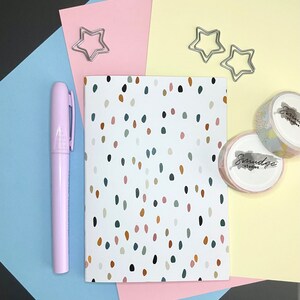 Puede incluir: Un cuaderno blanco con un patrón de confeti de colores. El cuaderno está sobre un fondo rosa, azul y amarillo con clips de papel en forma de estrella plateados.