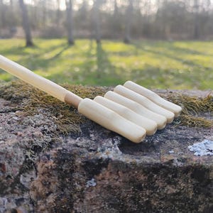 May include: A light-colored wooden back scratcher with a long handle and five rounded tines. The back scratcher is resting on a moss-covered rock, with a blurred forest background.