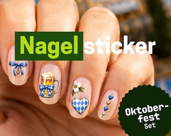 Op de afbeelding: Nagelstickers met Oktoberfest-thema aangebracht op vingernagels. De ontwerpen bevatten een blauwe strik, een bierpul met een blauw geblokte strik, een witte bloem, een blauw-wit geblokt hart en een rij kleine blauwe en gouden hartjes.