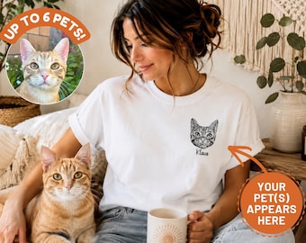 Puede incluir: Una camiseta blanca con una ilustración de líneas en blanco y negro de la cara de un gato en el lado izquierdo del pecho, con el nombre Klaus impreso en fuente cursiva debajo del gráfico. La camiseta tiene mangas cortas y cuello redondo.