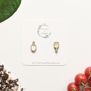 May include: A pair of wooden earrings featuring a small white shovel and a small green gardening fork. The earrings are on a white card with the text "The House of Marigolds" and "www.TheHouseofMarigolds.com".