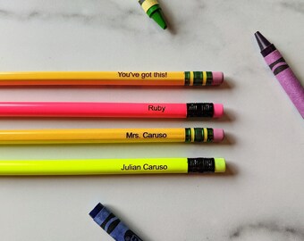 May include: Four personalised pencils in yellow, pink, and green with black erasers and text. The pencils have the following text: "You've got this!", "Ruby", "Mrs. Caruso", and "Julian Caruso". A purple and green crayon are also in the image.