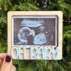 Puede incluir: Un marco de fotos de madera con una inscripción "OH BABY" en azul claro. El marco está diseñado para contener una imagen de ultrasonido.