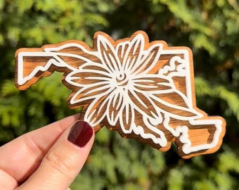 May include: A wooden cutout of the state of Maryland with a white floral design. The cutout is held in a hand with dark red nail polish.