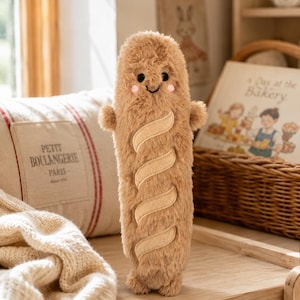 Può includere: Un peluche a forma di baguette con pelliccia sintetica marrone chiaro, piccoli occhi neri e guance ricamate in rosa. Il giocattolo presenta tre tagli diagonali in tessuto color crema sul davanti, piccole braccia arrotondate e una consistenza morbida.