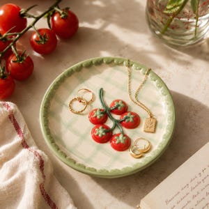 May include: A round ceramic trinket dish featuring a hand-painted light green plaid pattern and a scalloped edge. The center displays a raised, three-dimensional cluster of five red tomatoes on a green vine. Gold jewelry rests on the dish surface.