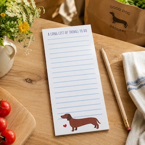 Può includere: Un blocco note rettangolare con sfondo bianco e linee orizzontali azzurre. Il testo in alto recita A LONG LIST OF THINGS TO DO in blu. In basso, è presente un'illustrazione di un bassotto marrone accanto a un piccolo cuore rosso.
