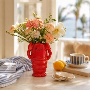 May include: A bright red ceramic lobster-shaped vase filled with a colourful bouquet of pink, peach, and white flowers. The vase features raised claw details and a simple smiling face. The arrangement sits on a surface with a blue and white striped cloth.