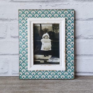 May include: A rectangular photo frame with a white inner frame and a patterned outer frame. The outer frame features a teal and white fan-shaped design. The frame holds a black and white photograph of a child. The text "sass & belle 6 x 10cm" is visible.