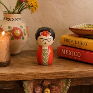 May include: A small, rounded ceramic figurine depicting a person with dark hair, a floral headpiece, and closed eyes. The figure wears a red garment with a green panel featuring a small white and pink flower pattern on the front of the torso.