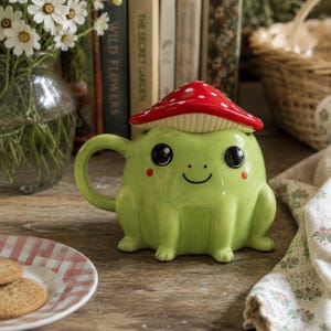 May include: A light green ceramic mug shaped like a frog with a handle on the left side. The mug features a removable lid designed as a red mushroom cap with white spots. The frog has large black eyes, a small smile, and two red circular cheek marks.