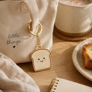 Op de afbeelding: Een goudkleurige metalen sleutelhanger met een crèmekleurige emaille bedel in de vorm van een sneetje brood met een lachend gezichtje, roze wangen en zwarte ogen. De bedel hangt aan een gouden ketting en ring aan een beige canvas tas.