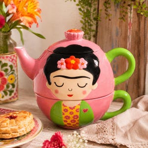 Könnte beinhalten: Ein stapelbares Set aus einer rosa Keramik-Teekanne und einer Teetasse mit dem gemalten Porträt einer Person mit schwarzem Haar, Blumenschmuck und geschlossenen Augen. Die Teekanne hat einen grünen Griff und Ausguss, während die Tasse eine grüne Jacke mit gelbem Blumenmuster und einem kleinen roten Herzdetail zeigt.