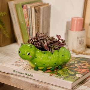 May include: A bright green ceramic caterpillar planter with a smiling face, pink cheeks, and white polka dots. The planter holds a small plant with variegated purple and green leaves, featuring a hollow opening on the back for the foliage.