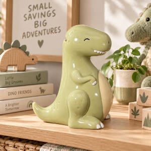 May include: A glossy, light green ceramic dinosaur coin bank with a smooth finish, a cream-colored belly, and a small coin slot on the top of its head. The dinosaur has a smiling expression with closed eyes and small white claws on its feet.