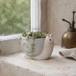 May include: A ceramic snail-shaped planter with a light blue shell and a cream-coloured body. The snail features two small antennae, black eyes, and a subtle pink cheek detail. A small green trailing plant is potted inside the shell portion.