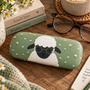 Peut inclure: Un étui à lunettes rigide rectangulaire de couleur vert sauge, orné d'une illustration centrale représentant un mouton blanc portant des lunettes rondes vertes, entouré d'un motif répétitif de petites fleurs blanches sur toute la surface.
