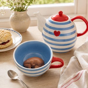 Puede incluir: Un juego de té de cerámica para una persona que incluye una tetera y una taza con rayas horizontales azules y blancas. La tetera tiene una tapa, un asa roja y un pequeño corazón rojo. La taza contiene una pequeña figura de perro de color marrón.