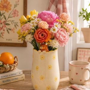 May include: A cream-coloured ceramic vase with a scalloped rim and a repeating yellow flower pattern. A small brown highland cow figurine is attached to the front rim. The vase is filled with a vibrant bouquet of pink, orange, yellow, and white flowers.
