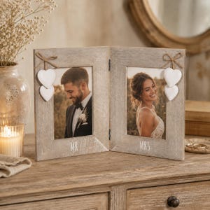 Può includere: Una cornice doppia incernierata in legno marrone chiaro con finitura anticata. La cornice sinistra presenta la scritta MR e due cuori bianchi appesi a un fiocco di juta. La cornice destra presenta la scritta MRS e due cuori bianchi.