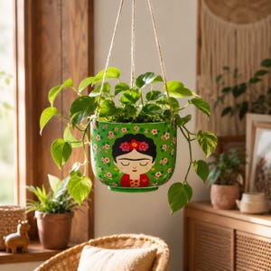 May include: A green hanging planter featuring a stylized illustration of a person with dark hair, a floral crown, and a red top. The planter is decorated with small pink and white flowers and is suspended by three thin ropes holding a green plant.