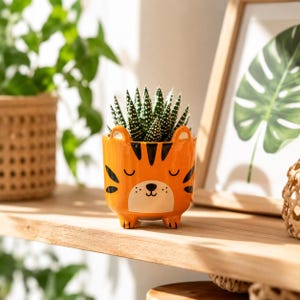 May include: A bright orange ceramic planter shaped like a tiger face with black stripes, small ears, and a white muzzle. The planter features a succulent plant with dark green, white-spotted leaves growing from the top and stands on four small feet.